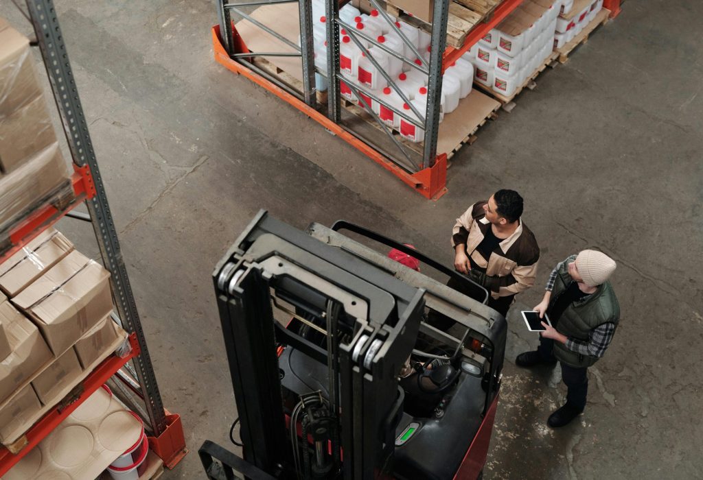 Two workers in a warehouse discussing logistics near a forklift captured from above.