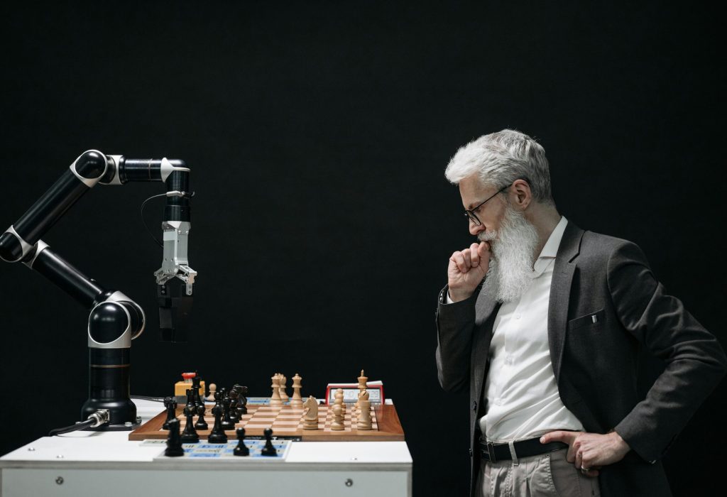 An elderly scientist contemplates a chess move against a robotic arm on a chessboard.